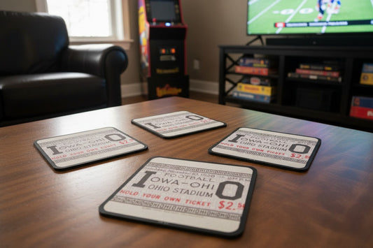Four coasters with 'Iowa Football vs. Ohio at Ohio Stadium' design on a wooden table in a game room with a television in background.