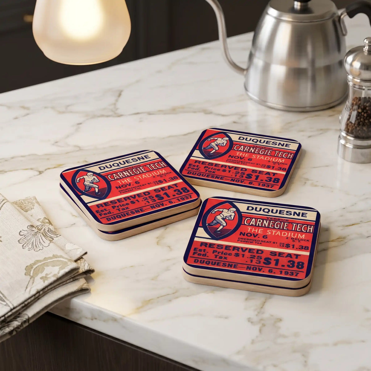 Square wooden ticket coasters with a vintage 1937 football ticket design for Duquesne Carnegie Tech are arranged on a white marble countertop next to a folded napkin, a silver kettle, and a pepper mill. Elements of image generated by AI