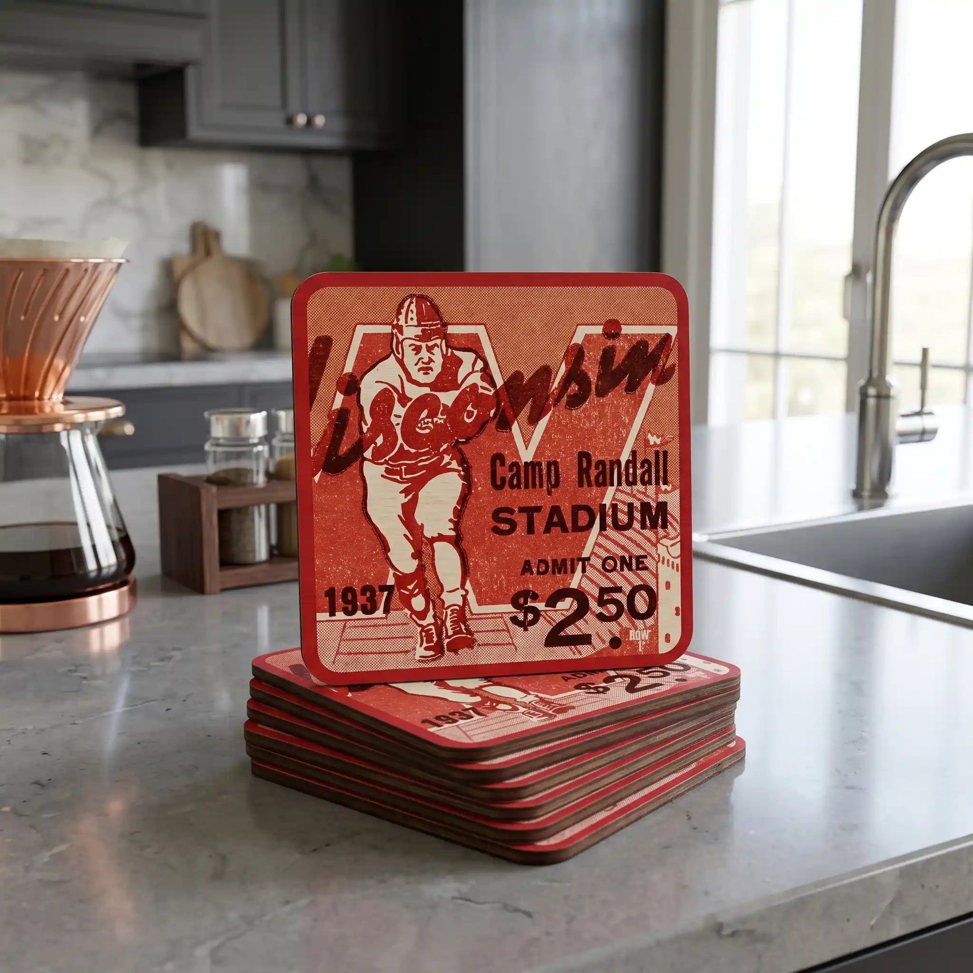 A stack of square red coasters with a vintage football stadium 1937 ticket design featuring the word Wisconsin and Camp Randall Stadium, displayed on a gray kitchen countertop.