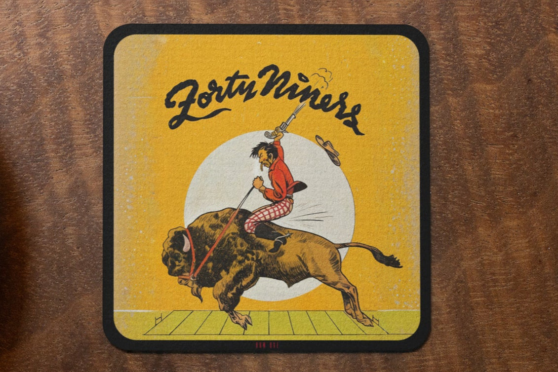 Row One Brand 1940s vintage football cover art birch wood coaster with 'Forty Niners' design featuring a person on a bison next to a cup of coffee and cookies on a wooden table.