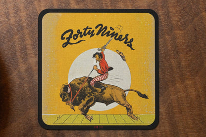 Row One Brand 1940s vintage football cover art birch wood coaster with 'Forty Niners' design featuring a person on a bison next to a cup of coffee and cookies on a wooden table.