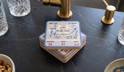 A stack of Row One Brand 1947 college football Army Note Dame ticket stub birch wood coasters on a dark marble countertop, surrounded by kitchen items like a gold faucet, glass cups, and a soap dispenser