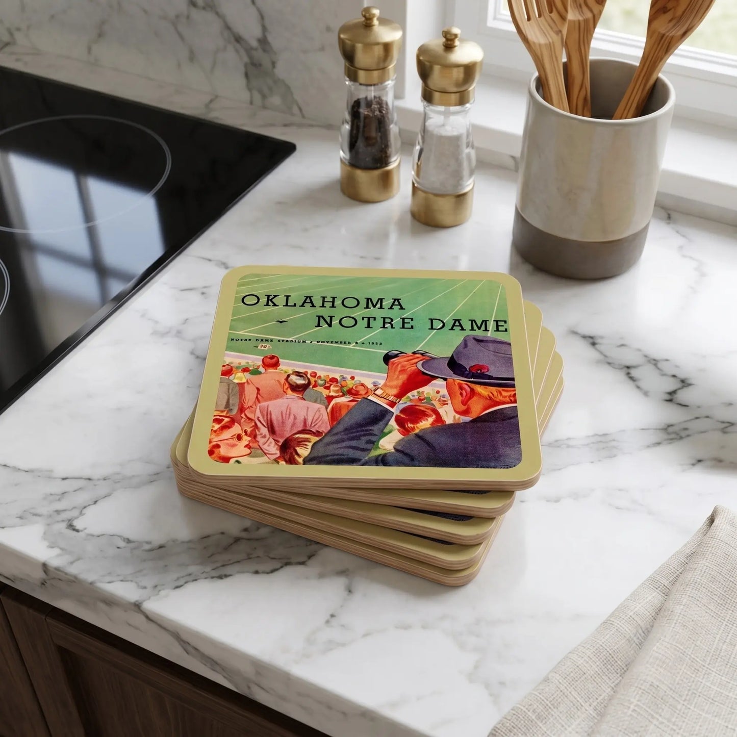 A stack of square coasters with rounded corners sits on a white marble kitchen countertop. The coasters have a gold border and a printed design featuring Oklahoma Notre Dame football players in a stadium, with the text 'OKLAHOMA NOTRE DAME' at the top, date of game, and vintage football stadium art.