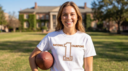 A smiling woman stands outdoors on a grassy field holding a football, wearing a white Row One Brand vintage college football T-shirt with orange brown text that reads 'National Champions 1963' and a large number 1 graphic.