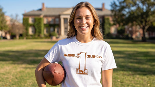 A smiling woman stands outdoors on a grassy field holding a football, wearing a white Row One Brand vintage college football T-shirt with orange brown text that reads 'National Champions 1963' and a large number 1 graphic.