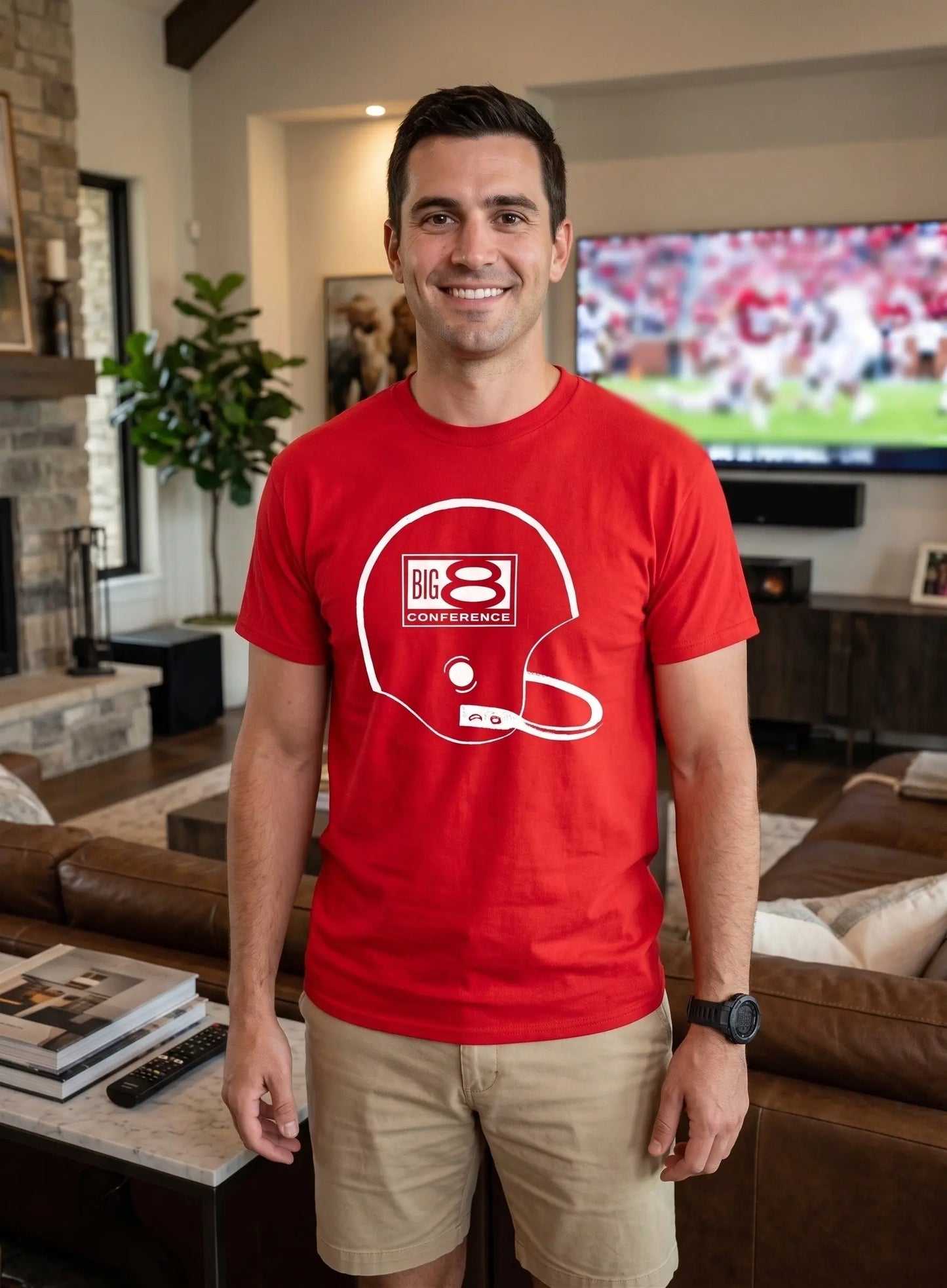 A man wearing a bright red crewneck t-shirt with a white graphic of a football helmet and 'BIG 8 CONFERENCE' text, standing in a modern living room with a TV in the background.