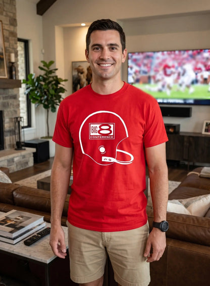 A man wearing a bright red crewneck t-shirt with a white graphic of a football helmet and 'BIG 8 CONFERENCE' text, standing in a modern living room with a TV in the background.