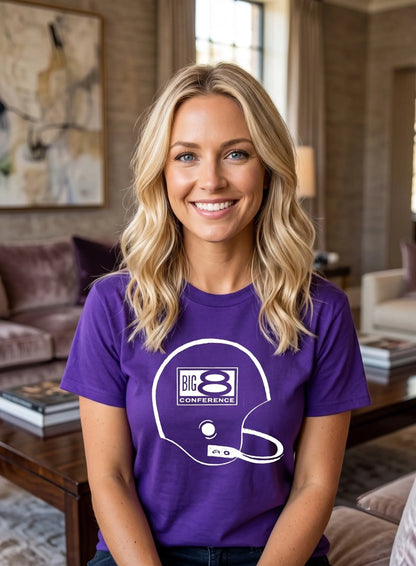 A smiling blonde woman wearing a Row One Brand purple t-shirt with a white graphic of a football helmet and the text 'BIG 8 CONFERENCE' posed in a cozy living room setting.