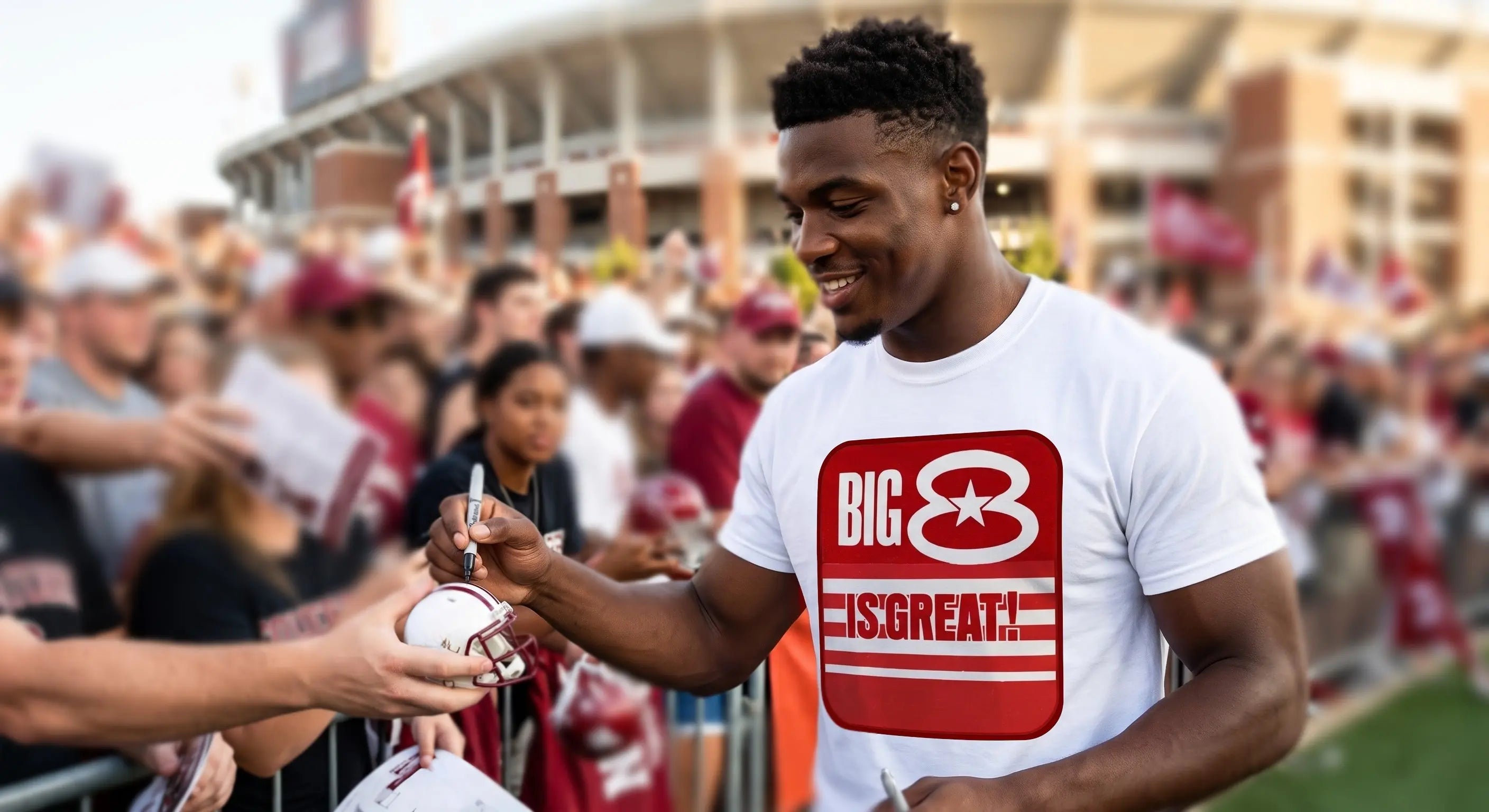 A man in a Row One Brand white graphic t-shirt with a red and white 'BIG 8 IS GREAT!' logo autographs a football helmet at a stadium event, with fans and stadium buildings blurred in the background.