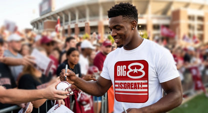 A man in a Row One Brand white graphic t-shirt with a red and white 'BIG 8 IS GREAT!' logo autographs a football helmet at a stadium event, with fans and stadium buildings blurred in the background.