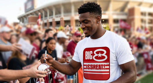 A man in a Row One Brand white graphic t-shirt with a red and white 'BIG 8 IS GREAT!' logo autographs a football helmet at a stadium event, with fans and stadium buildings blurred in the background.