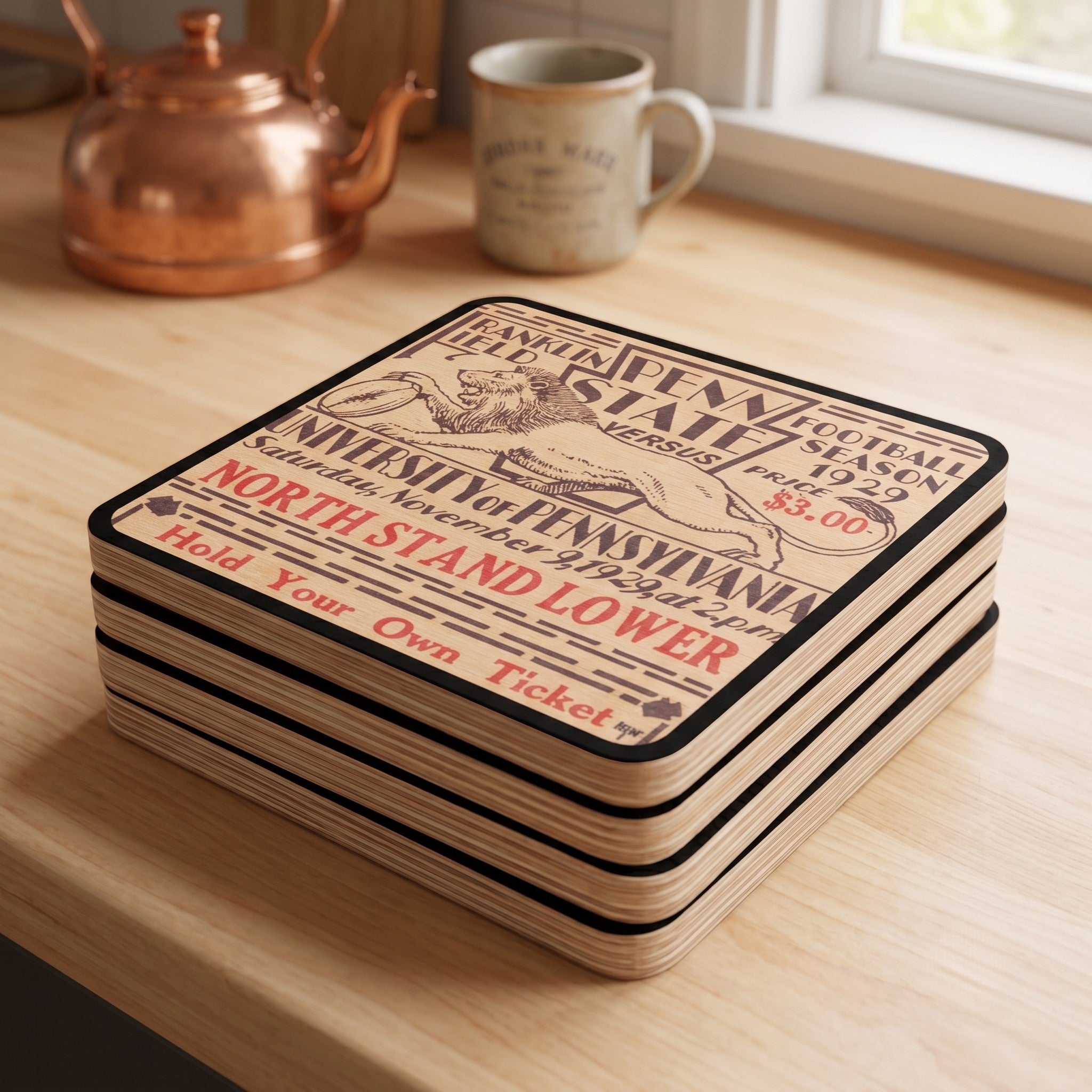A stack of 4 square wooden coasters with Row 1 trademark in lower right corner with a vintage public domain historic college football ticket 1929 Penn State at Penn football ticket design, displayed on a light wood table near a window with a copper kettle and mug in the background.