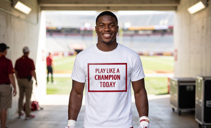 A smiling man stands in a stadium tunnel wearing a white graphic T-shirt with a red border and the text 'PLAY LIKE A CHAMPION TODAY' printed on the front.