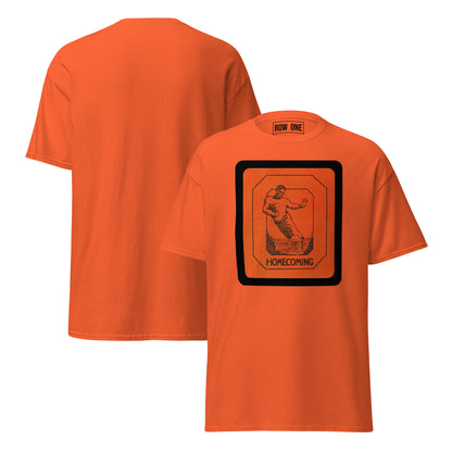An orange short-sleeve t-shirt shown from both front and back. The front features a large black and orange graphic on the chest depicting a vintage football player in motion inside a bordered square, with the word 'HOMECOMING' printed below.