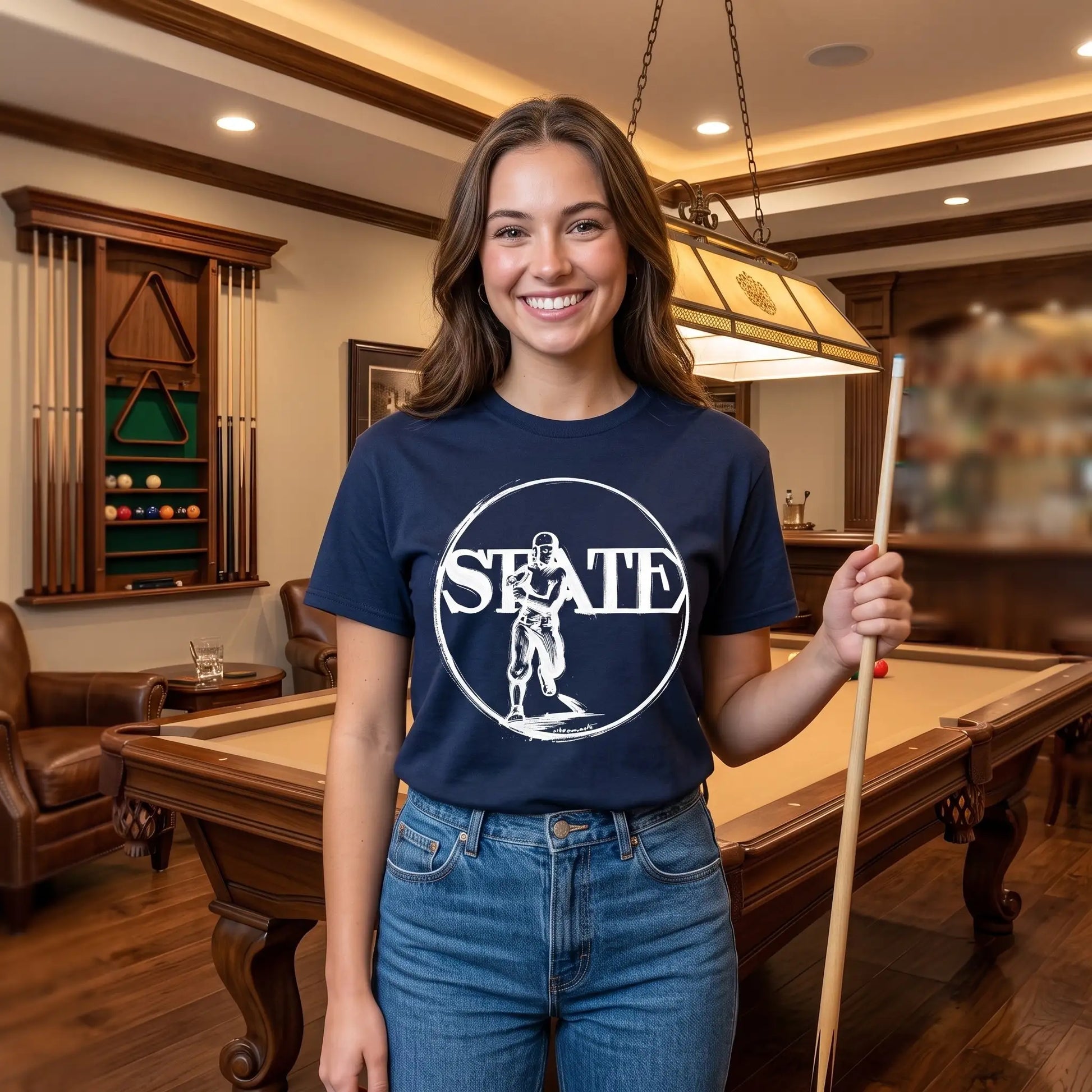 A woman stands in a pool room wearing a navy blue graphic t-shirt with a white circular logo that reads 'STATE' and features a football player illustration from early 1900s. She is holding a pool cue stick and wearing blue jeans.