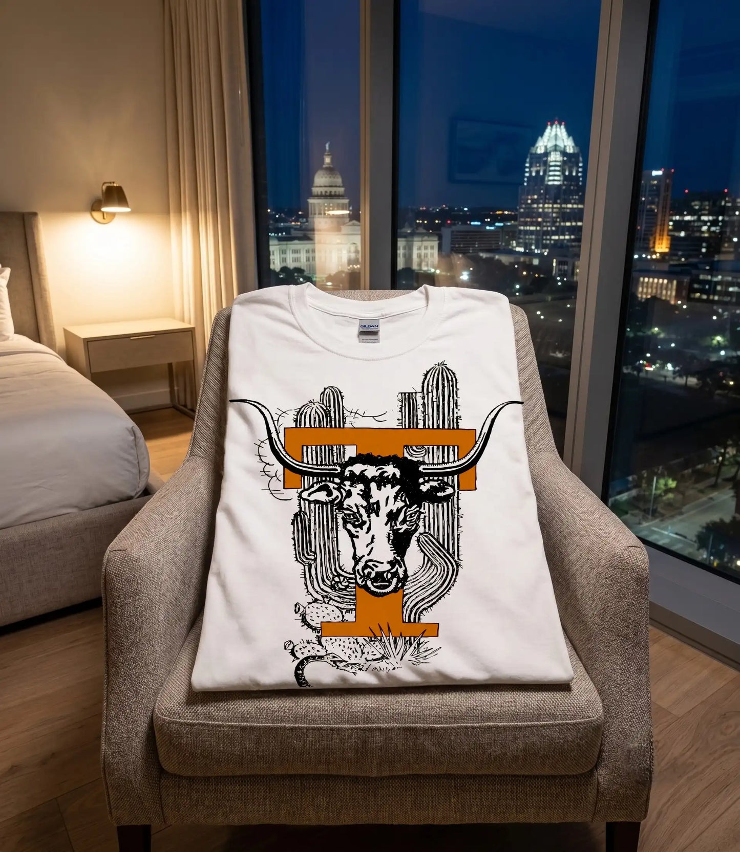 A white crewneck t-shirt with a graphic of a longhorn steer and cacti, styled on a beige armchair in a modern bedroom with a city skyline at night visible through the window.