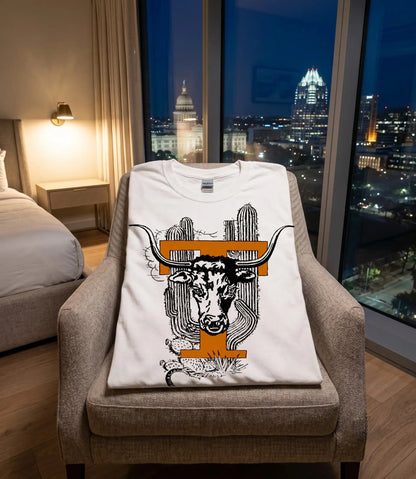 A white crewneck t-shirt with a graphic of a longhorn steer and cacti, styled on a beige armchair in a modern bedroom with a city skyline at night visible through the window.